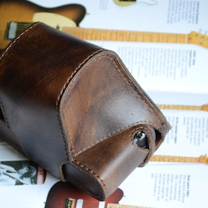 Op de afbeelding: Een donkerbruine lederen cameratas met zichtbare stiksels langs de randen. De tas heeft een gestructureerd, hoekig ontwerp met een klepsluiting en een kleine ronde opening aan de zijkant, afgewerkt met een gladde, gepolijste textuur.