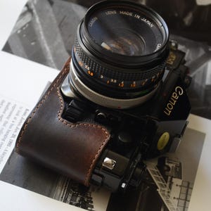 May include: A black Canon camera with a lens made in Japan, featuring a brown leather case. The camera is resting on a page with black and white images and text.
