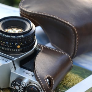 Op de afbeelding: Een zilverkleurige Pentax K1000 filmcamera met een zwarte SMC Pentax-A lens bevestigd. De camera is gedeeltelijk omhuld door een donkerbruine lederen beschermhoes met zichtbare witte stiksels langs de randen. De lens toont diafragma- en focusmarkeringen.