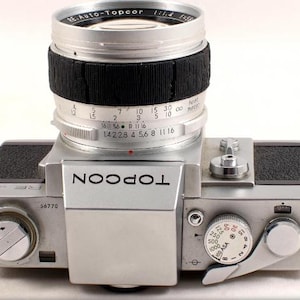 May include: A silver and black 35mm film camera with a silver lens. The lens has a black rubber ring around it and the text "RE AUTO-Topcor lild 1250" printed on it. The camera has the text "TOPCOR" printed on the front in black letters.