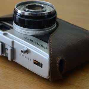 以下が含まれることがあります： シルバーの金属製ボディと黒いレンズ鏡筒を持つオリンパスTrip 35フィルムカメラ。ダークブラウンのレザー製保護ケースに部分的に収められている。上面にはOLYMPUS TRIP 35の文字、シリアルナンバー1895829、小さなオレンジ色のインジケーターが表示されている。