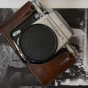 May include: A silver Pentacon Six TL camera with a black lens cap, in a brown leather case. The camera is on top of a black and white photo.