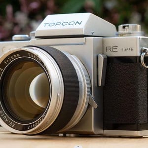 Può includere: Una macchina fotografica a pellicola da 35 mm di colore argento e nero con un obiettivo Topcon RE Super. L'obiettivo ha una lunghezza focale di 58 mm e un'apertura massima di f/1.4.