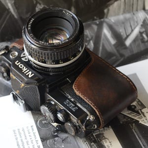 May include: A black Nikon FM2 film camera with a brown leather case and a black lens. The lens is labeled "Nikon" and "50mm". The camera is resting on a book with black and white images.