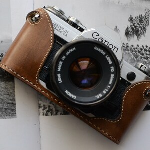 Könnte beinhalten: Eine silberne Canon AE-1 Kamera mit einer braunen Ledertasche. Die Kamera hat ein Canon FD 50mm 1:1.8 S.C. Objektiv. Die Kamera liegt auf einer Seite aus einem Buch mit einem Schwarzweißfoto einer Gruppe von Menschen.