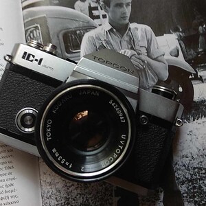 Può includere: Una macchina fotografica a pellicola da 35 mm Topcon IC-1 Auto di colore argento e nero con un obiettivo Tokyo Kogaku 1:5.3 53 mm. La macchina fotografica è appoggiata su un libro con una foto in bianco e nero di un uomo in un'auto.