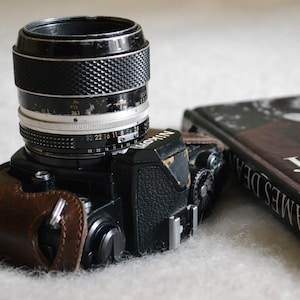 Puede incluir: Una cámara de película Nikon negra con un objetivo plateado y negro, que incluye una funda de cuero marrón en el lateral y un libro titulado James Dean parcialmente visible a la derecha del cuerpo de la cámara sobre una superficie blanca.