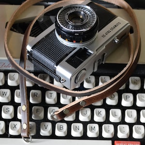 Peut inclure: Un appareil photo Olympus Trip 35 avec une sangle en cuir est posé sur une machine à écrire Brother Deluxe. L'appareil photo est argenté et noir, avec un objectif proéminent. La machine à écrire a des touches blanches et une touche "TAB" rouge.