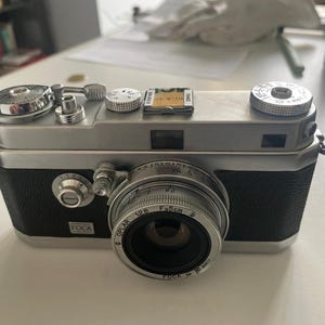 May include: A vintage silver and black FOCA camera with a leather strap. The camera has a lens with the text "OPLAR 1:2.8 F=5cm" and a film card on top. The camera is a classic piece of photography equipment.