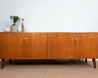 Könnte beinhalten: Ein Mid-Century-Modern-Holzschrank mit vier Türen und abgewinkelten Beinen. Der Schrank hat eine warme, mittelbraune Farbe mit sichtbarer Holzmaserung. Dekorative Gegenstände, darunter eine kleine Pflanze, stehen oben. Der Schrank ist etwa 152 cm breit.