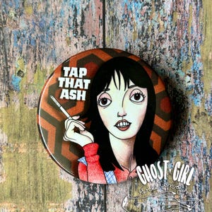 May include: A round button with a cartoon illustration of a woman holding a cigarette. The button has the text "TAP THAT ASH" in white block letters. The background is a brown and orange geometric pattern.