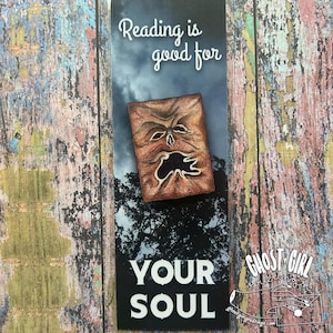 May include: A bookmark with the text "Reading is good for YOUR SOUL." The bookmark features a detailed illustration of a brown, weathered book with a face on the cover. The background has a dark, cloudy sky and a silhouetted tree line.
