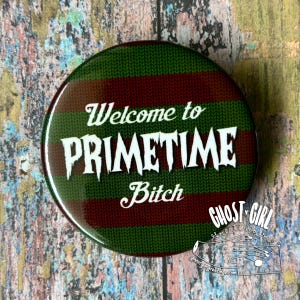 May include: Round button with a green and burgundy sweater pattern background. The white text reads "Welcome to PRIMETIME Bitch". The button has a glossy finish and a white border. The Ghost Girl Greetings logo is in the lower right corner.