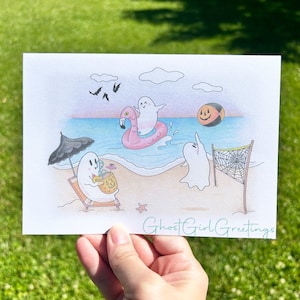 May include: A hand-drawn illustration of three ghosts enjoying a day at the beach. One ghost is riding a pink flamingo float, another is playing volleyball, and the third is sitting in a beach chair with a pumpkin-shaped drink. The illustration is on a white background with the text "Ghost Girl Greetings" at the bottom.