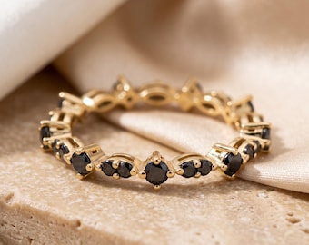 Op de afbeelding: Een gouden eeuwigheidsring met een doorlopende rij ronde zwarte stenen in een klauwzetting. De ring heeft een gepolijste metalen afwerking en een uniform ontwerp dat de hele band omcirkelt, met een reeks donkere, ronde edelstenen.