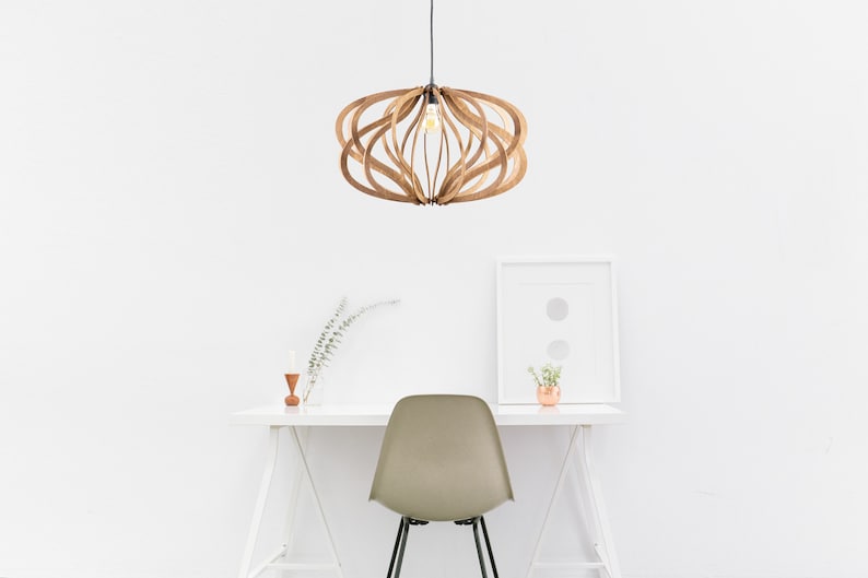 Op de afbeelding: Een houten hanglamp met een uniek, geometrisch ontwerp hangt aan het plafond boven een wit bureau met een stoel. De lamp is gemaakt van dunne, gebogen stukken hout die een ronde vorm cre&euml;ren. De lamp wordt verlicht door een enkele gloeilamp.