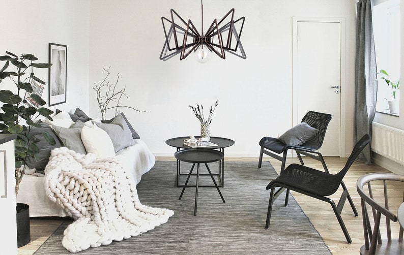 May include: A modern living room with a black geometric chandelier, a white couch with a chunky knit throw blanket, two black mesh chairs, and a set of black nesting coffee tables.