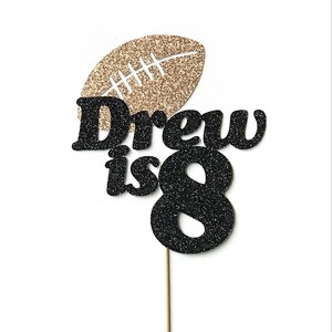 May include: Black and gold glitter cake topper with the text "Drew is 8" and a football design.