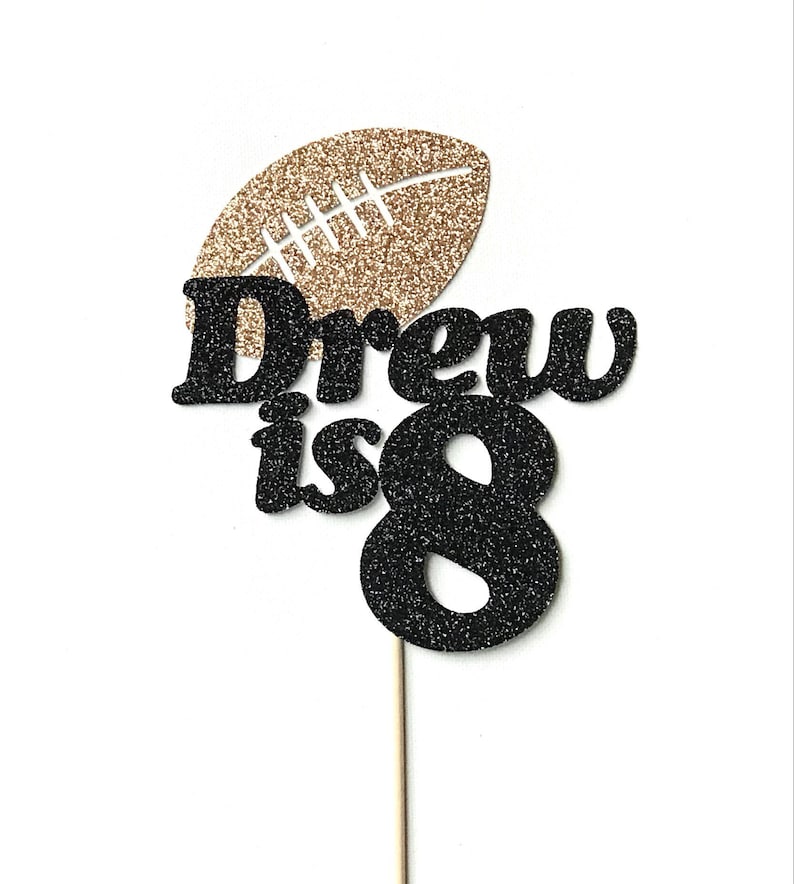 May include: Black and gold glitter cake topper with the text "Drew is 8" and a football design.