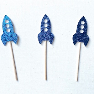 May include: Three blue glitter rocket cupcake toppers on wooden sticks. The rockets have three white circles for windows.