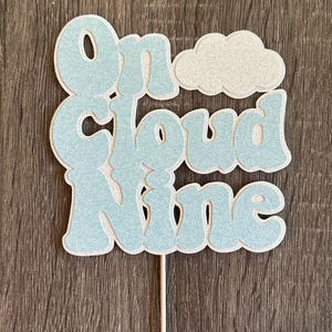 May include: A cake topper featuring the text On Cloud Nine in light blue glittery letters with a white outline. A glittery white cloud shape is positioned above the word Cloud. The entire design is attached to a thin wooden stick at the bottom.
