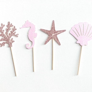 May include: Four pink and gold glitter sea-themed cake toppers. The toppers include a seahorse, a starfish, a seashell, and coral.