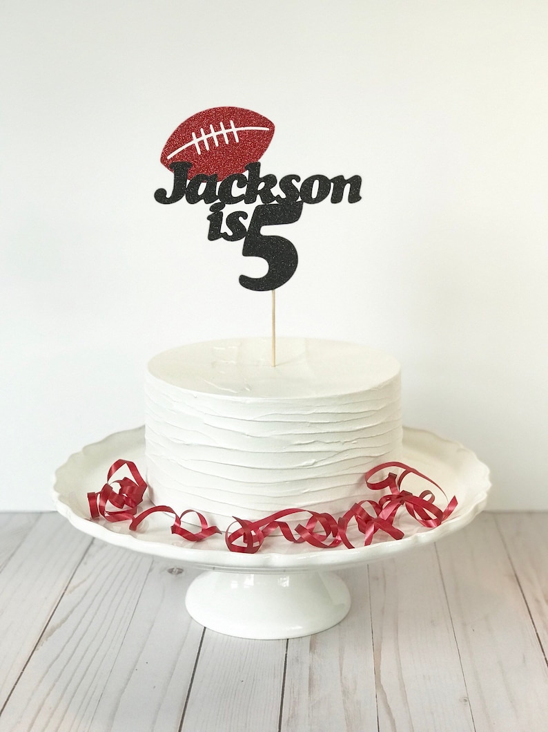 May include: A white cake with a red and black glitter cake topper that says "Jackson is 5" with a red glitter football on top.