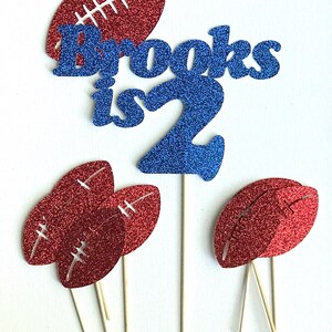 May include: A set of cake toppers featuring a blue glitter "Brooks is 2" and red glitter footballs. The footballs are on sticks and can be used to decorate cupcakes or a cake.