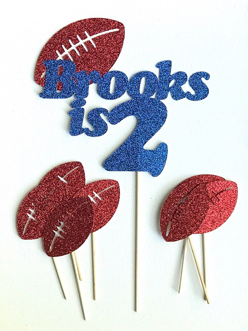 May include: A set of cake toppers featuring a blue glitter "Brooks is 2" and red glitter footballs. The footballs are on sticks and can be used to decorate cupcakes or a cake.