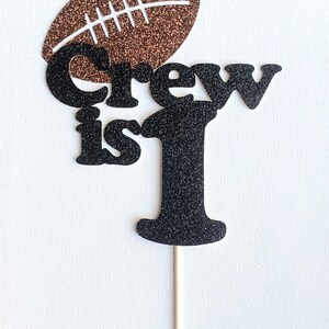 May include: Black and brown glitter cake topper with the words "Crew is 1" and a football on top.