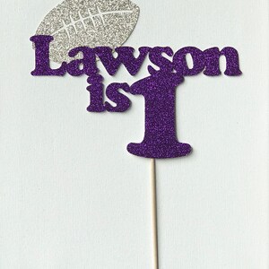 May include: Purple glitter cake topper with the text "Lawson is 1" and a silver glitter football.