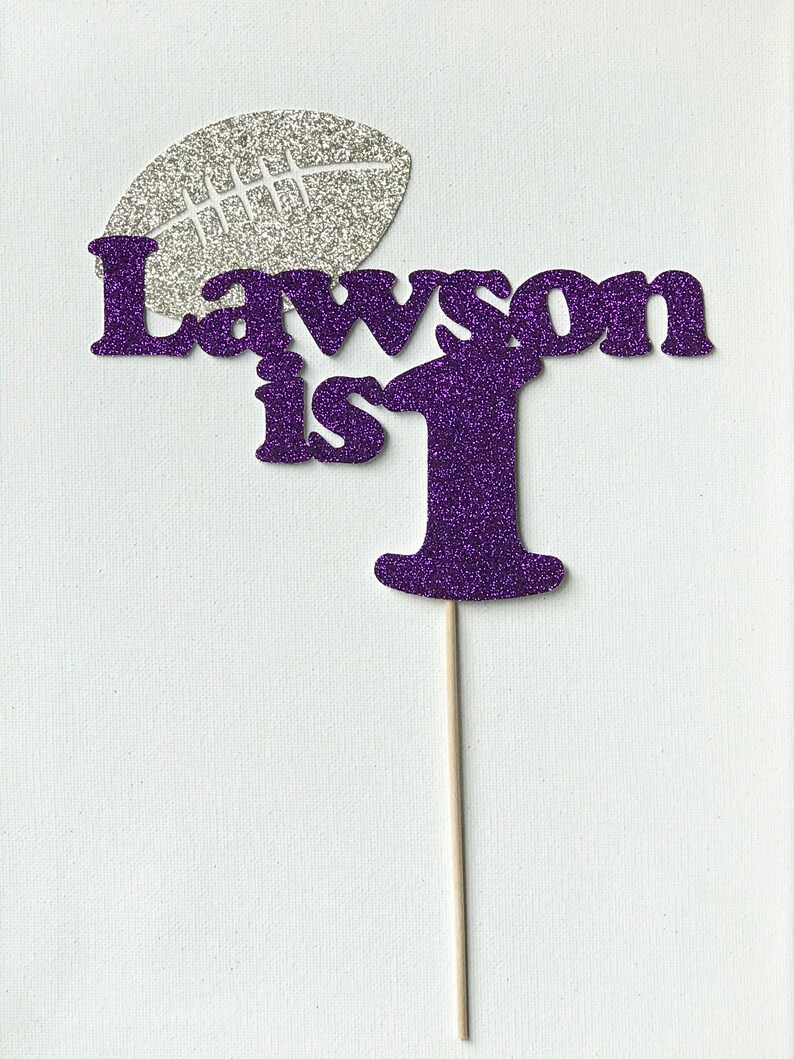 May include: Purple glitter cake topper with the text "Lawson is 1" and a silver glitter football.