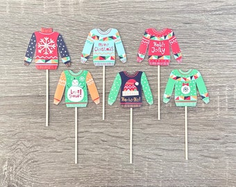 May include: Six festive Christmas sweater cupcake toppers. The toppers are printed with various holiday designs and sayings, including "Merry Christmas", "Let It Snow!" and "Ho-ho-ho!"