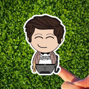 May include: A sticker featuring a cartoon character with brown hair and a smiling face. The character wears a blazer, bow tie, and pants. The shirt reads "Seattle Mariners Baseball Club." The sticker is held by a finger against a green leafy background.