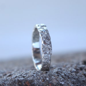 May include: A silver ring with a textured, rough surface. The ring is standing upright on a gray, textured surface.