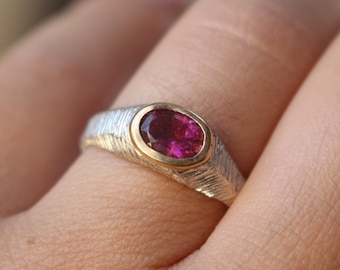 Könnte beinhalten: Ein silberner Ring mit einer strukturierten Band und einer goldenen Fassung. Der Ring hat einen rosafarbenen ovalen Edelstein.