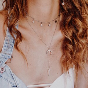 May include: A woman wearing a white tank top with a denim overall dress. She is wearing a silver necklace with a crescent moon pendant and a silver choker necklace with star charms.