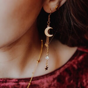 May include: Gold crescent moon earrings with a dangling chain featuring small stars and a clear crystal. The earrings are worn by a person with gold glitter makeup.
