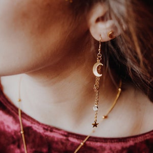 May include: Gold crescent moon dangle earrings with a star charm and a small crystal bead. The earrings are hanging from a person's ear.