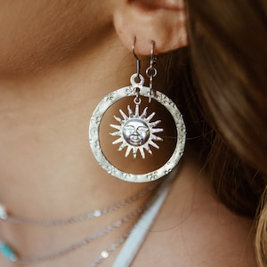 May include: Silver hoop earrings with a sun face design. The earrings are hanging from a person's ear.