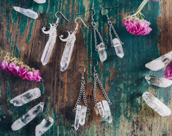 May include: A pair of silver earrings with clear quartz crystals hanging from a silver chain. The earrings are on a wooden surface with other clear quartz crystals.