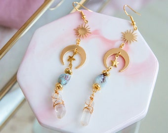 May include: Gold dangle earrings with a crescent moon, star, and sun charm. The earrings also feature a clear quartz crystal and a blue and white agate bead.
