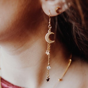 May include: Gold crescent moon dangle earrings with a small star charm and a clear crystal bead. The earrings are on a model's ear.