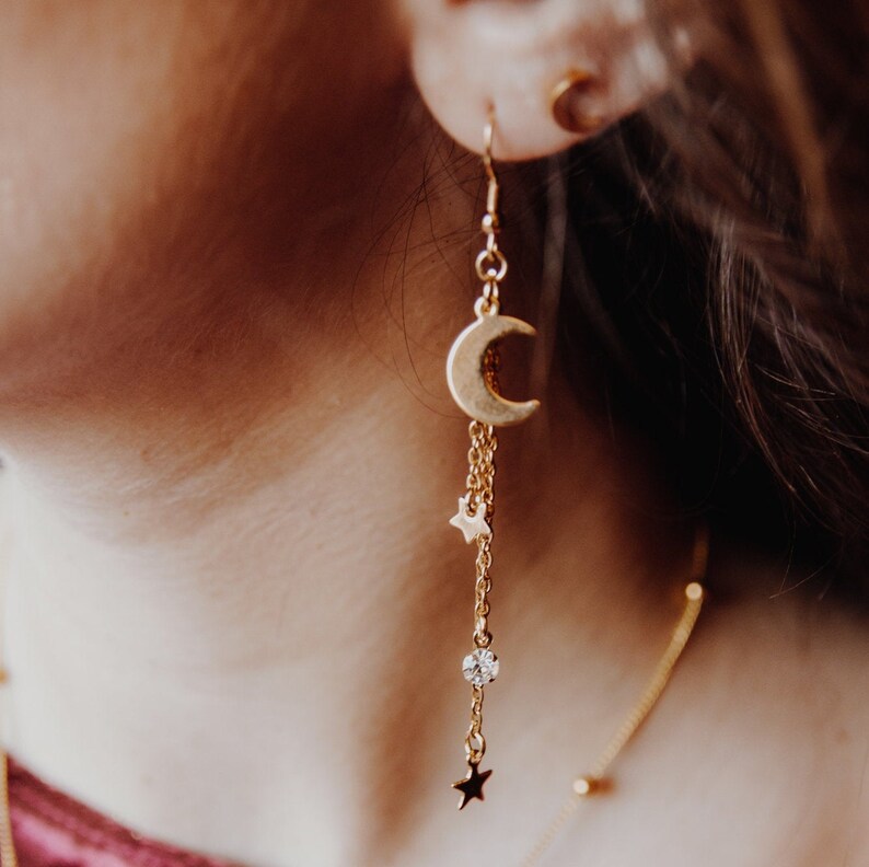May include: Gold crescent moon dangle earrings with a small star charm and a clear crystal bead. The earrings are on a model's ear.