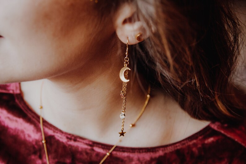 May include: Gold crescent moon dangle earrings with a star charm and a small crystal bead. The earrings are hanging from a person's ear.