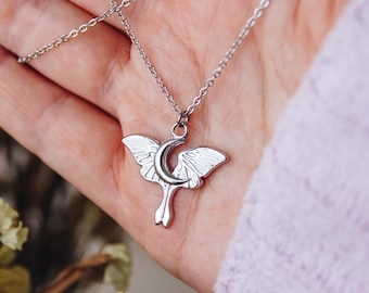 Op de afbeelding: Een zilveren ketting met een hanger in de vorm van een wassende maan en een nachtvlinder. De hanger is gedetailleerd met delicate vleugels en een wassende maanvorm.