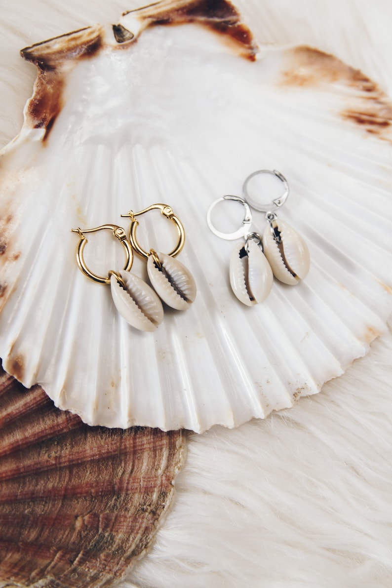 May include: Two pairs of earrings with gold and silver hoops and cowrie shell charms. The earrings are displayed on a white seashell.