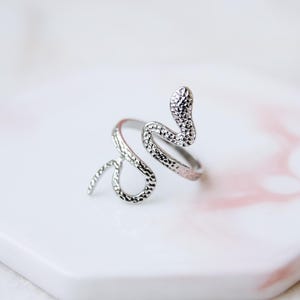 May include: A silver snake ring with a textured finish. The ring is shaped like a snake, with its head raised. The ring sits on a white and pink hexagonal surface.