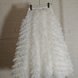 Peut inclure: Une jupe blanche composée de multiples couches horizontales de tissu tulle à volants. La jupe possède une taille élastiquée et est suspendue à un cintre en plastique transparent. Le modèle présente des rangées denses de tulle blanc.