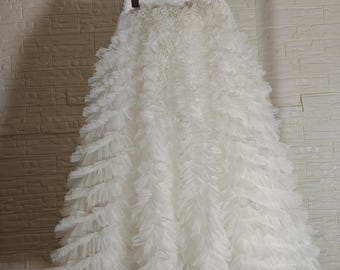 May include: A white, layered skirt with a ruffled design, hanging from a black hanger. The skirt is made of a soft, flowing fabric, creating a voluminous silhouette. The skirt is a solid white color.
